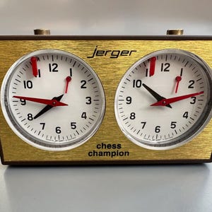 May include: A vintage Jerger chess clock with a gold-colored face and two round clock faces. The clocks have white backgrounds, black numbers, and red hands. The text "Jerger" is above the clocks, and "chess champion" is below.
