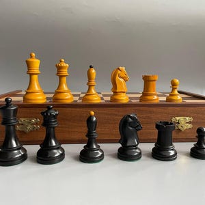 以下が含まれることがあります： 木製のチェスセット。黒と黄色の駒が木製の盤上に配置されています。駒は木製の箱の中にあります。駒には、キング、クイーン、ビショップ、ナイト、ルーク、ポーンが含まれています。