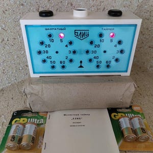 May include: A vintage chess timer with a white and blue face, featuring time indicators and the word "BLITZ". The timer has two black knobs and a light-up display. Includes batteries and a manual.
