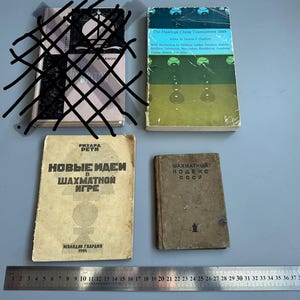 May include: Four vintage chess books are displayed on a grey surface. The books have aged covers with titles in English and Russian. A metal ruler with centimetre measurements is at the bottom of the image.