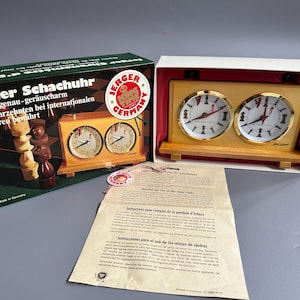 May include: A vintage Jerger Schachur chess clock in its original box. The clock has two round faces with black Roman numerals and red hands. The box is green and black with German text and a "King Time Germany" sticker.