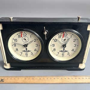 Puede incluir: Un reloj de ajedrez vintage negro y crema con dos esferas circulares. Cada esfera tiene números negros, un segundero rojo y un pequeño logotipo. Una regla de madera está en la parte inferior.