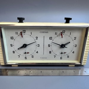 May include: Vintage ivory-colored chess clock with two clock faces. Each clock face has black hands, black numbers, and red second hands. The clock is labeled "CORSAR" and "4 steine". A metal ruler is placed below the clock.
