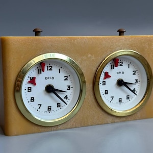 May include: A vintage chess clock with a light brown case and two circular white clock faces. Each face has black hands, gold-coloured rims, and black numerals. The clock has two small brown knobs on top.