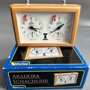 Könnte beinhalten: Schachuhr aus Holz mit zwei weißen Zifferblättern, schwarzen Zeigern und roten Markierungen. Das Zifferblatt trägt die Aufschrift "ARADORA Classic" und "JEWELLED". Die Uhr steht auf einer blauen Schachtel mit der Aufschrift "ARADORA SCHACHUHR".