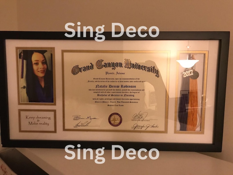 May include: A framed diploma from Grand Canyon University for Natalie Denise Robinson, with a graduation tassel in purple and orange. The diploma is in a black frame with a white mat. The frame has a small photo of a woman in a cap and gown, and the text "Keep dreaming & Make reality" below the photo.