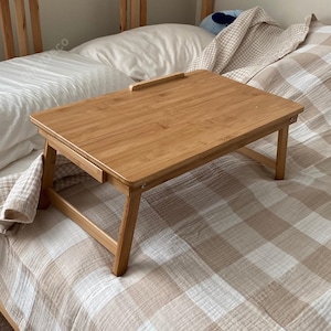 May include: A light brown bamboo bed tray with folding legs. The tray has a rectangular surface with a raised edge and a slot for a tablet or phone. The tray is on a bed with a white pillow and a beige and white checkered blanket.