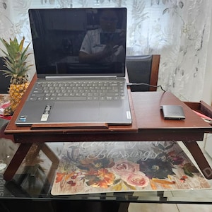 May include: A dark brown wooden laptop desk with a pull-out drawer and angled legs. A gray laptop is on the desk, along with a small, silver device. A pineapple sits nearby. The desk is on a glass table.