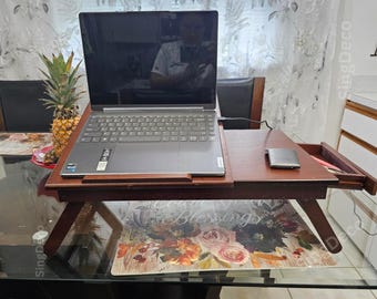 May include: A dark brown wooden laptop desk with a pull-out drawer and angled legs. A gray laptop is on the desk, along with a small, silver device. A pineapple sits nearby. The desk is on a glass table.