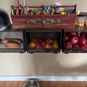 May include: A wall-mounted storage unit with three wire baskets, a wooden toolbox, and two glass jars. The baskets hold sweet potatoes and apples. A blue pencil sharpener sits on the left. The toolbox is filled with pencils and pens.