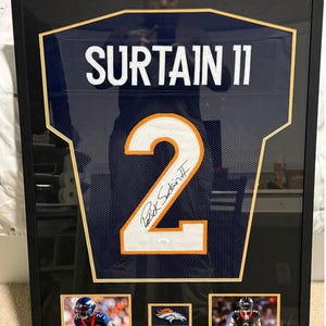 May include: Framed Denver Broncos jersey with the name "SURTAIN 11" and the number "2" prominently displayed. The jersey is signed and framed with two action photos and a team logo. The frame is black.