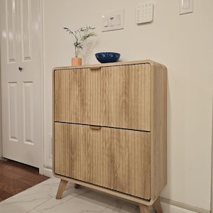 May include: A light-colored wooden cabinet with two drawers and a ribbed front design. The cabinet has a mid-century modern style with angled legs. A small blue bowl and a potted plant sit on top.