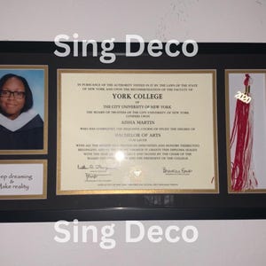 May include: A framed diploma from York College of the City University of New York, awarded to Aisha Martin for a Bachelor of Arts degree. The diploma is decorated with a gold tassel and a small photo of the graduate. The frame has a gold border and a black background. The text "Keep dreaming & Make reality" is printed on a small plaque below the photo.