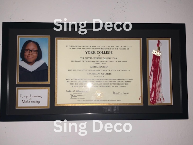 May include: A framed diploma from York College of the City University of New York, awarded to Aisha Martin for a Bachelor of Arts degree. The diploma is decorated with a gold tassel and a small photo of the graduate. The frame has a gold border and a black background. The text "Keep dreaming & Make reality" is printed on a small plaque below the photo.