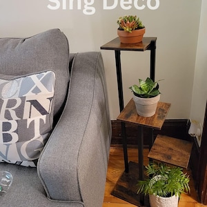 May include: A black metal plant stand with three tiers. Each tier is made of wood with a brown finish. The stand is decorated with three potted plants, including a succulent, a snake plant, and a green leafy plant. The stand is in a living room setting next to a gray couch. The text "Sing Deco" is visible in the image.