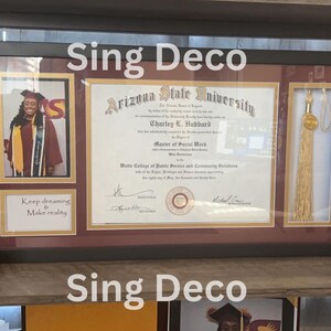 May include: A framed diploma from Arizona State University for a Master of Social Work degree. The diploma is in a black frame with a maroon mat. There is a photo of a woman in a maroon graduation gown and a gold tassel in the frame. The text "Sing Deco" is written at the top and bottom of the frame.