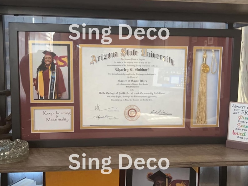 May include: A framed diploma from Arizona State University for a Master of Social Work degree. The diploma is in a black frame with a maroon mat. There is a photo of a woman in a maroon graduation gown and a gold tassel in the frame. The text "Sing Deco" is written at the top and bottom of the frame.