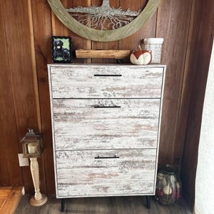 May include: A white and brown distressed wood-look chest of drawers with three drawers and black handles. The chest is set against a wood-paneled wall, with decorative items on top.
