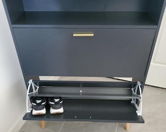 May include: A dark gray shoe cabinet with a gold-colored handle. The cabinet has a top shelf and a drop-down door revealing a shoe rack. Two pairs of sneakers are visible inside the cabinet. The cabinet is supported by short wooden legs.