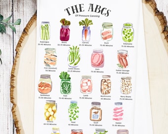 May include: White kitchen towel with a colorful illustration of the ABCs of Pressure Canning. The towel features various canned goods with their respective canning times. The towel is displayed on a wooden surface with greenery and cooking utensils.
