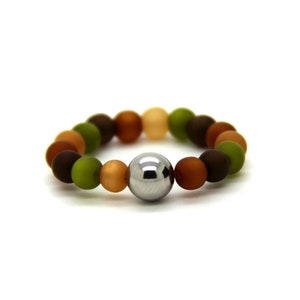 Op de afbeelding: Een bruin, groen en oranje kralenarmband met een zilveren metalen kraal in het midden.