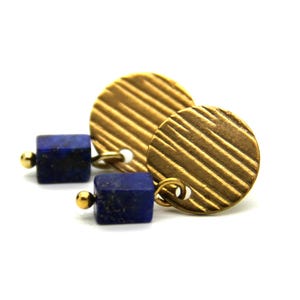 May include: A pair of earrings featuring gold-colored, ridged circular pendants and dark blue, cube-shaped lapis lazuli stones. The earrings have small gold-colored ball studs. The jewelry is set against a white background.