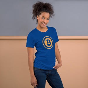 May include: A blue t-shirt featuring a Bitcoin symbol in a gold circle. The Bitcoin symbol is a stylized letter "B" with vertical lines. The shirt is worn by a person with dark hair and blue jeans.