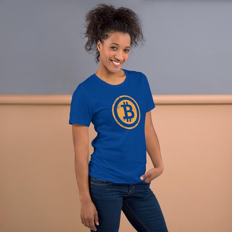 May include: A blue t-shirt featuring a Bitcoin symbol in a gold circle. The Bitcoin symbol is a stylized letter "B" with vertical lines. The shirt is worn by a person with dark hair and blue jeans.