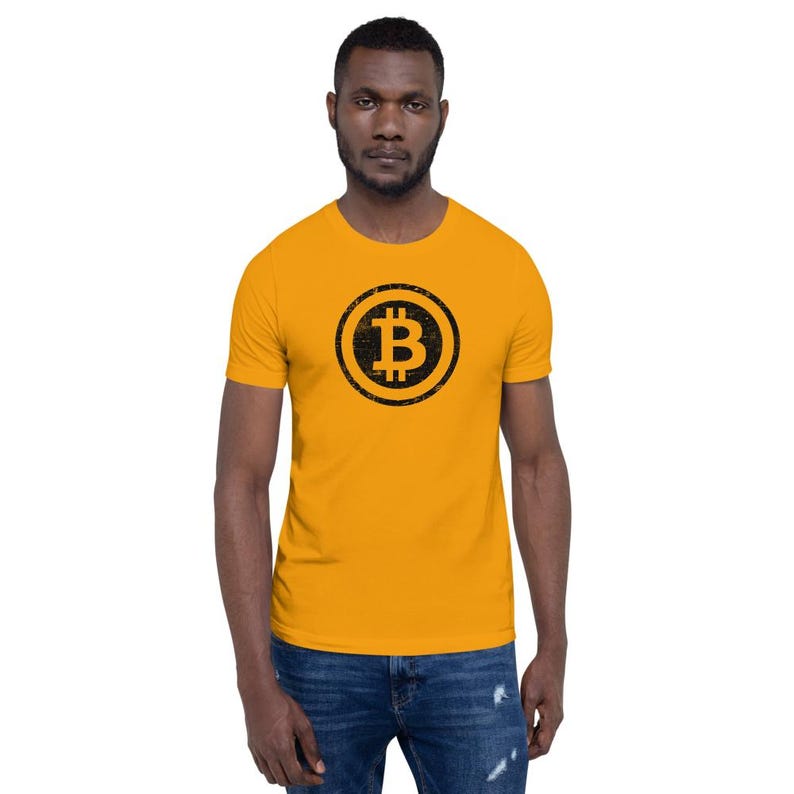 May include: Mustard yellow t-shirt featuring a black Bitcoin symbol within a circle. The Bitcoin logo is slightly distressed. The shirt is short-sleeved and worn by a person in blue jeans.