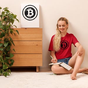 May include: A red t-shirt with a black Bitcoin symbol is worn by a person sitting on a white rug. A matching Bitcoin symbol is on a white canvas leaning against a wooden dresser. A green plant is to the left of the dresser.