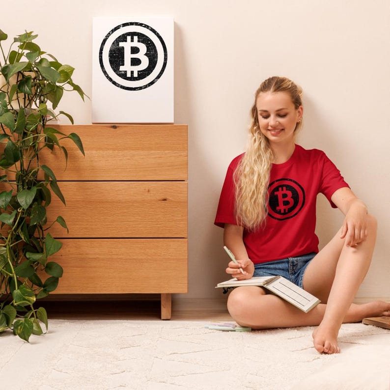 May include: A red t-shirt with a black Bitcoin symbol is worn by a person sitting on a white rug. A matching Bitcoin symbol is on a white canvas leaning against a wooden dresser. A green plant is to the left of the dresser.