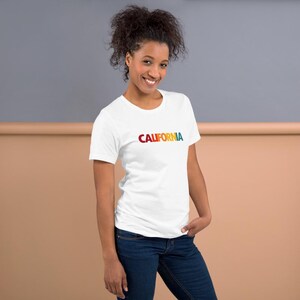 May include: White t-shirt with the word "CALIFORNIA" printed across the chest in a rainbow gradient. The shirt is paired with blue jeans. The model is smiling.