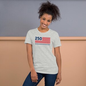 K&ouml;nnte beinhalten: Hellgraues T-Shirt mit einem grafischen Motiv einer stilisierten amerikanischen Flagge und der Zahl "250" in Blau. Das Flaggen-Design umfasst rote Streifen, blaue Sterne und die Zahl "250". Das Shirt ist aus weichem Material.