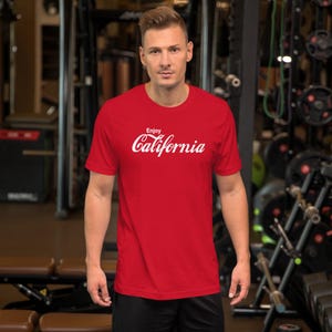 May include: A red t-shirt with the words "Enjoy California" in white script. The shirt is short-sleeved and appears to be made of a soft material. The person wearing the shirt is standing in a gym.