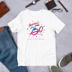 May include: White t-shirt with a patriotic design. The shirt features the text "America 250 1776-2026" in red, white, and blue, surrounded by stars. The phrase "Years in the Making" is printed below the main text.