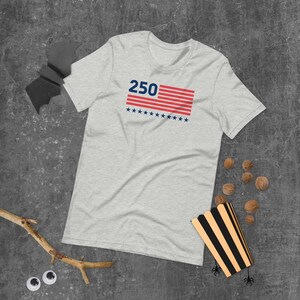 K&ouml;nnte beinhalten: Hellgraues T-Shirt mit der Zahl "250" &uuml;ber einem rot-wei&szlig; gestreiften Design mit blauen Sternen. Das kurz&auml;rmelige T-Shirt ist aus einem weichen Material. Das T-Shirt liegt auf einer grauen Oberfl&auml;che.