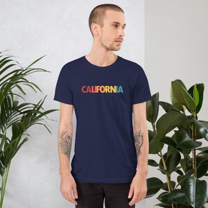 May include: Navy blue t-shirt with the word "CALIFORNIA" printed across the chest in a retro rainbow gradient. The shirt is a short-sleeved crew neck style, perfect for casual wear. The design is simple and eye-catching.