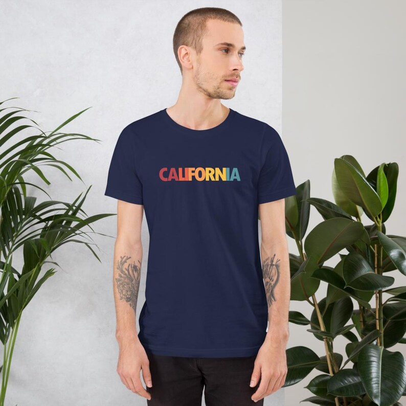 May include: Navy blue t-shirt with the word "CALIFORNIA" printed across the chest in a retro rainbow gradient. The shirt is a short-sleeved crew neck style, perfect for casual wear. The design is simple and eye-catching.