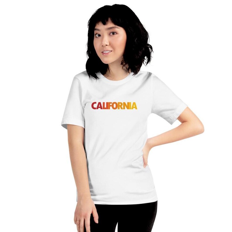 Op de afbeelding: Wit T-shirt met het woord "CALIFORNIA" in een rood-gele gradi&euml;nt. De top met korte mouwen wordt gedragen door een persoon met zwart haar. Het T-shirt is een casual stijl.