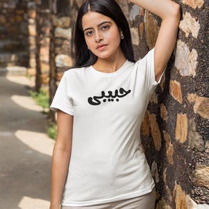 K&ouml;nnte beinhalten: Wei&szlig;es T-Shirt mit dem arabischen Wort &bdquo;Habibi&ldquo; in Schwarz aufgedruckt. Das T-Shirt hat einen Rundhalsausschnitt und wird von einer Person getragen. Die Person lehnt an einer Steinmauer. Der Hintergrund umfasst einen Steinbogen.