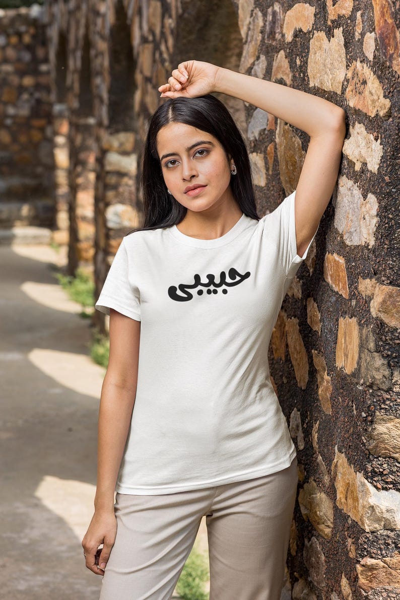 K&ouml;nnte beinhalten: Wei&szlig;es T-Shirt mit dem arabischen Wort &bdquo;Habibi&ldquo; in Schwarz aufgedruckt. Das T-Shirt hat einen Rundhalsausschnitt und wird von einer Person getragen. Die Person lehnt an einer Steinmauer. Der Hintergrund umfasst einen Steinbogen.