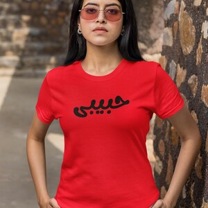 K&ouml;nnte beinhalten: Ein rotes T-Shirt mit Rundhalsausschnitt und schwarzer arabischer Schrift auf der Brust. Das Shirt hat kurze &Auml;rmel und wird getragen. Die Person tr&auml;gt eine helle Hose und eine Sonnenbrille.