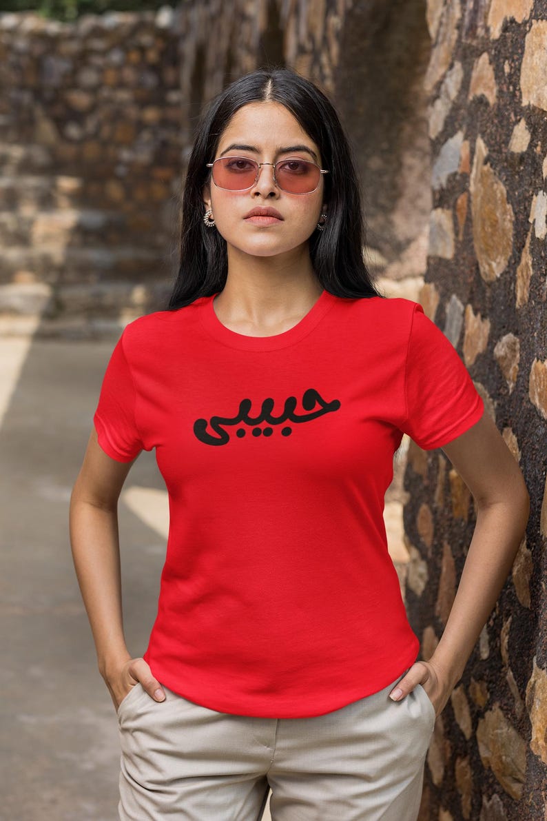 K&ouml;nnte beinhalten: Ein rotes T-Shirt mit Rundhalsausschnitt und schwarzer arabischer Schrift auf der Brust. Das Shirt hat kurze &Auml;rmel und wird getragen. Die Person tr&auml;gt eine helle Hose und eine Sonnenbrille.