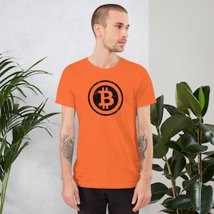 May include: Orange short-sleeve t-shirt featuring a black Bitcoin symbol in a circular design. The Bitcoin symbol is a stylized letter "B" with vertical lines. The shirt is made of a soft material.
