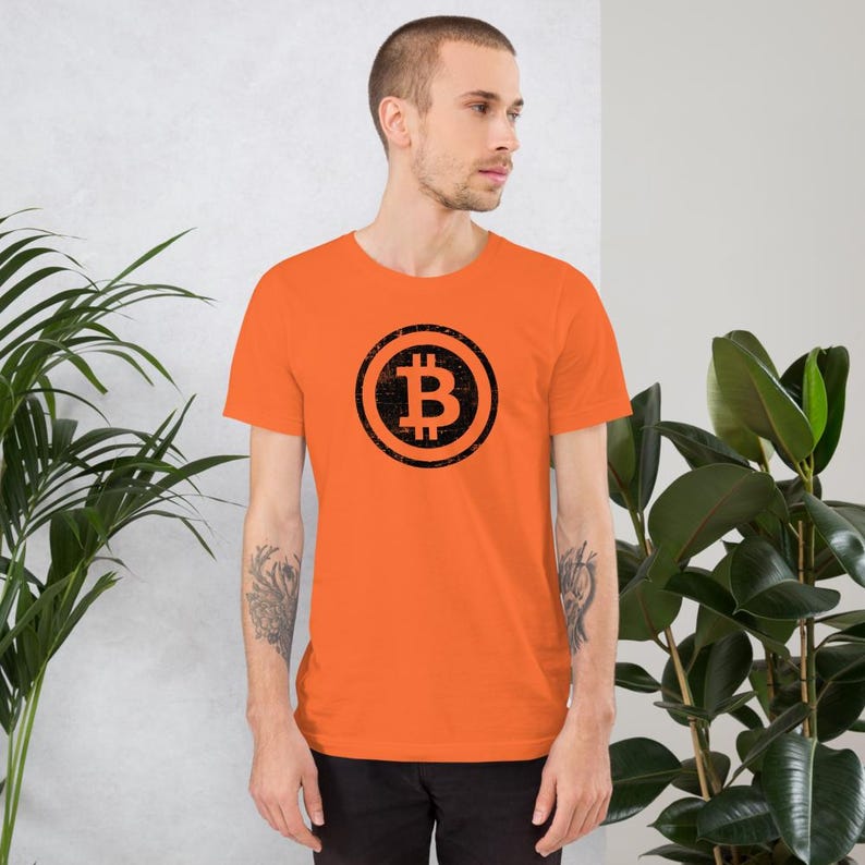 May include: Orange short-sleeve t-shirt featuring a black Bitcoin symbol in a circular design. The Bitcoin symbol is a stylized letter "B" with vertical lines. The shirt is made of a soft material.
