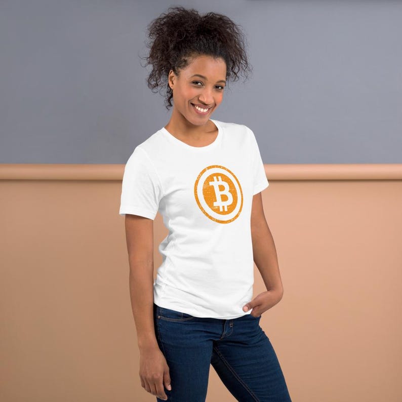 May include: White t-shirt with a round orange Bitcoin symbol graphic. The Bitcoin symbol features a stylized letter "B" inside a circle. The shirt is made of a soft material and is paired with blue jeans.