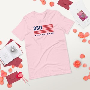 K&ouml;nnte beinhalten: Ein hellrosa T-Shirt mit der Zahl "250" &uuml;ber einem Design, das an die amerikanische Flagge erinnert. Die Flagge hat rote und wei&szlig;e Streifen und blaue Sterne. Das T-Shirt ist aus weichem Stoff.