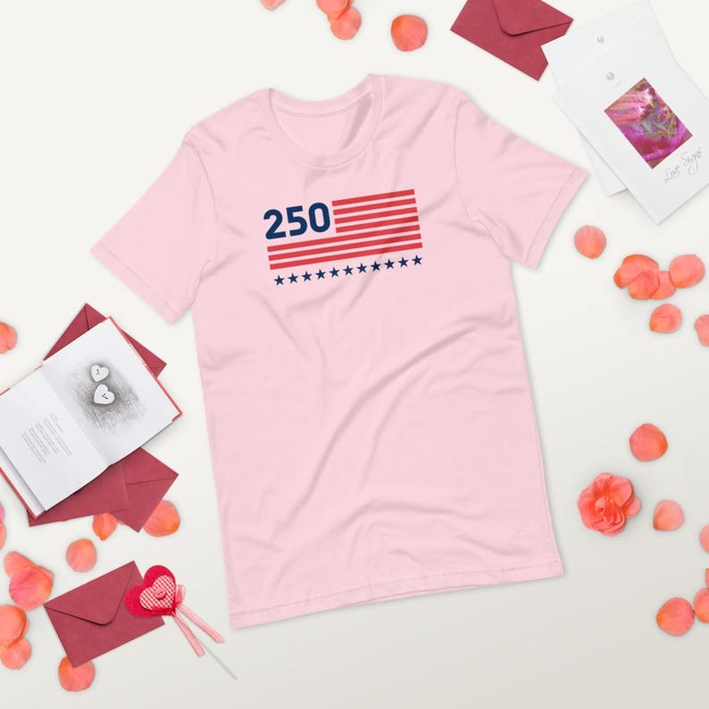 K&ouml;nnte beinhalten: Ein hellrosa T-Shirt mit der Zahl "250" &uuml;ber einem Design, das an die amerikanische Flagge erinnert. Die Flagge hat rote und wei&szlig;e Streifen und blaue Sterne. Das T-Shirt ist aus weichem Stoff.