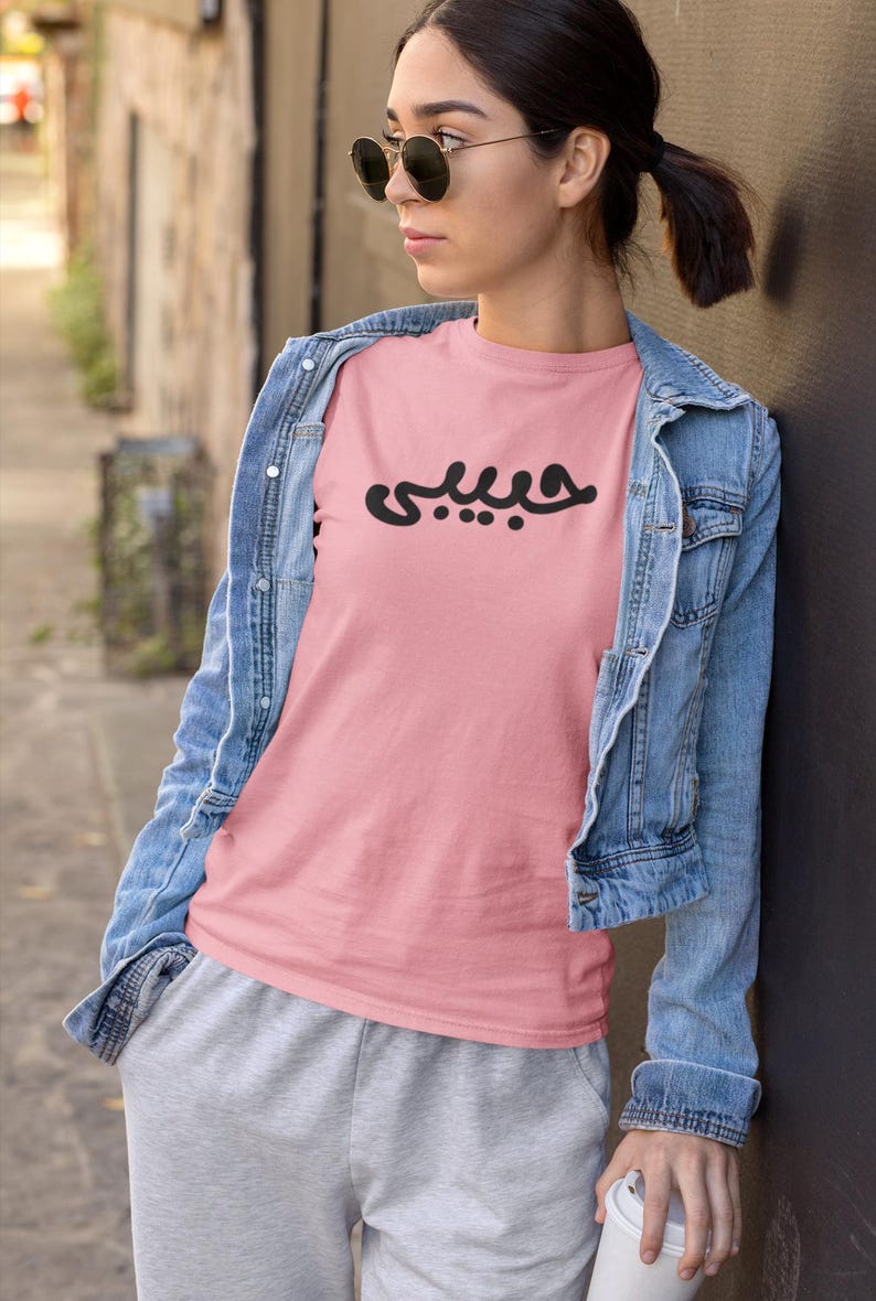 K&ouml;nnte beinhalten: Hellrosa T-Shirt mit dem arabischen Wort &bdquo;Habibi&ldquo; in schwarzer Schrift. Das Shirt wird mit einer Jeansjacke und grauer Jogginghose getragen. Die Person tr&auml;gt eine Sonnenbrille.