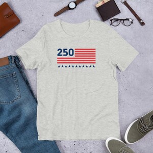 K&ouml;nnte beinhalten: Ein hellgraues T-Shirt mit einem grafischen Design. Das Design zeigt die Zahl "250" &uuml;ber einem rot-wei&szlig; gestreiften Rechteck und eine Reihe blauer Sterne darunter. Das T-Shirt ist aus einem weichen Material gefertigt.